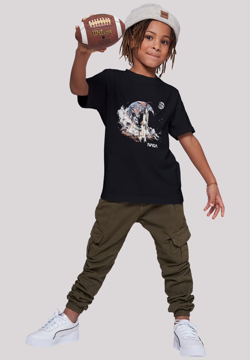 Schwarzes NASA-Grafik-T-Shirt mit Raumfähren-Design, olivfarbene Cargo-Hosen und weiße Sneakers. Das Kind hält einen braun-weißen Fußball.