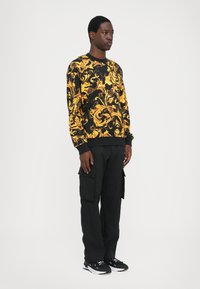 Must mustam sweatshirt, mille keerukate kuldsete lillemustritega, koos mustade cargo-pükste ja mustade spordijalatsitega, millel on valged aktsendid.