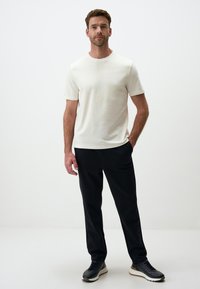 Leichtes, kurzärmeliges, cremefarbenes Hemd für Herren kombiniert mit schwarzer Hose und schwarzen Sneakers, auf schlichtem weißen Hintergrund.