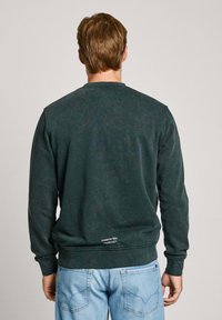 Sudadera verde oscuro con cuello redondo, puños y dobladillo acanalados, con un pequeño logo en la parte baja de la espalda. Llevada con jeans de color azul claro.