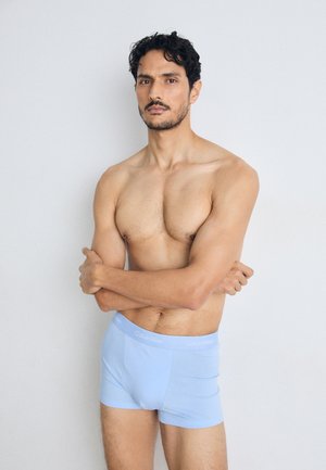 Boxers en coton bleu clair avec logo Calvin Klein sur la taille, présentant un design ajusté et une texture lisse. Le modèle pose les bras croisés.