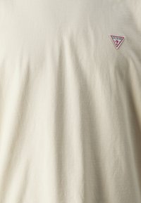 Tricou din bumbac bej, cu textură netedă și guler rotund. Prezintă un logo brodat triangular cu "GUESS" în roșu și albastru.