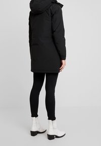 Černá péřová bunda s kapucí, dlouhého střihu. Spárov ána s černými legínami a bílými kotníkovými botami s vroubkovanými detaily a blokovými podpatky.