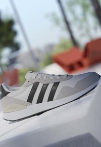 Baskets Adidas blanches et grises avec des bandes noires, placées à l'extérieur sur une surface blanche, avec un arrière-plan vert et marron flou.