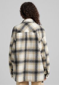 Persona con cabello rizado que lleva una sobrecamisa de cuadros en beige, negro y blanco, de pie de espaldas contra un fondo liso gris claro.