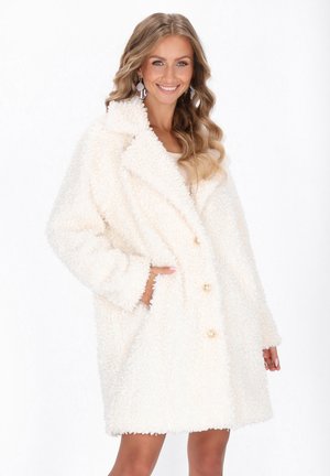 Woman smiling, wearing a white textured teddy coat with gold buttons, hand in pocket, wavy blonde hair, and iridescent earrings.