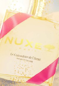 Calendario dell'Avvento NUXE Paris, in vetro trasparente con dettagli dorati, nastro rosa, contiene particelle brillanti e presenta testo in rilievo.