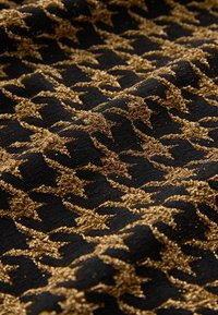 Crni materijal s teksturiranim zlatnim houndstooth uzorkom, s blistavim koncima koji stvaraju sjajni efekt na površini.
