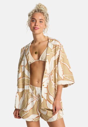 Giovane donna con capelli biondi ricci indossa una camicia oversize beige e bianca a fiori, shorts abbinati e un top bikini crema all'uncinetto.