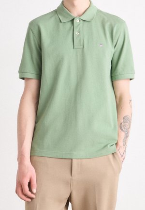 Homme portant un polo à manches courtes vert clair et un pantalon beige, avec la main droite dans la poche et un tatouage sur l'avant-bras gauche.