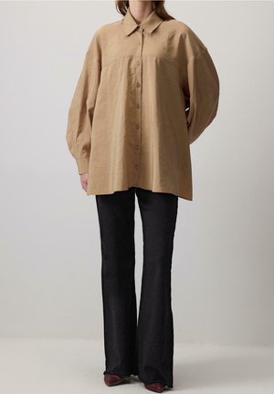 Kvinde iført oversized beige skjorte med lange ballonærmer, kombineret med sorte vide bukser og mørke spidse sko.