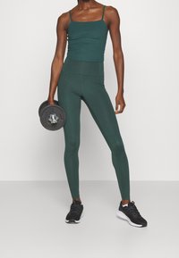 Grünes, geripptes Tanktop mit dünnen Trägern, kombiniert mit hochgeschnittenen grünen Leggings. Schwarze Sneakers und ein schwarzes Gewichte in der Hand.