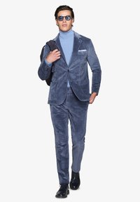 Blå kordfloret-kostym med en texturerad polokrage, slim-fit byxor och en blazer. Accessoarer inkluderar solglasögon och en svart axelväska.
