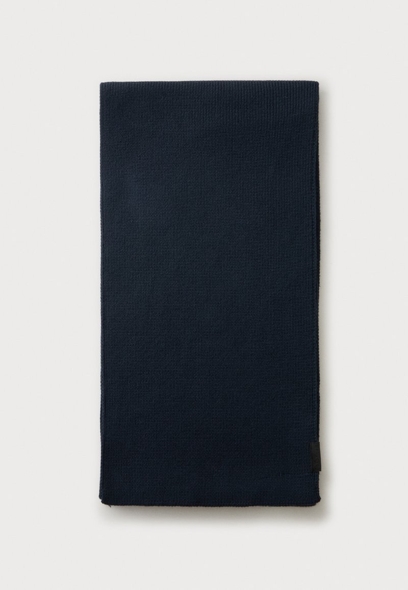 Écharpe tricotée bleu marine foncé pliée sur un fond clair uni avec une petite étiquette noire sur le bord.