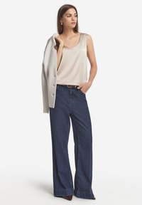 Beige satin ärmlös topp, vida blå denimbyxor, ljusgrå cardigan hängande över axeln, brun bälte och mörka skor.