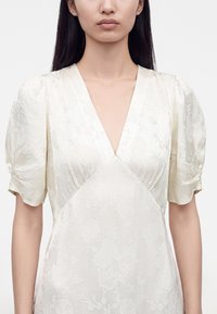 Femme portant une robe blanche à texture florale avec des manches courtes bouffantes et un profond décolleté en V, montrée des épaules à la taille.