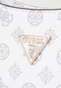 Bijela torba teksture sivi uzorak loga. Sadrži zlatni metalni logo u obliku trokuta s natpisom "GUESS USA" i detaljem u obliku upitnika.
