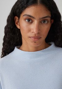 Hellblaues strukturiertes Oberteil mit hohem Kragen und feinem Muster. Der Stoff wirkt weich und leicht im Design.