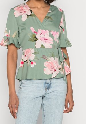 Femme portant un chemisier cache-cœur vert sauge avec un imprimé floral rose et des manches courtes à volants, associé à un jean bleu clair.