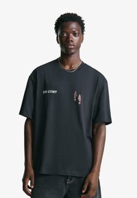 Svart oversized t-shirt i bomull med texten "TOY STORY" och en design med skosulor i brunt. Kortärmad, enkel rund halsringning.