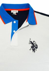 Polo blanco con cuello azul y rojo, dos botones blancos y logotipo bordado en negro de un jugador de polo en el pecho.