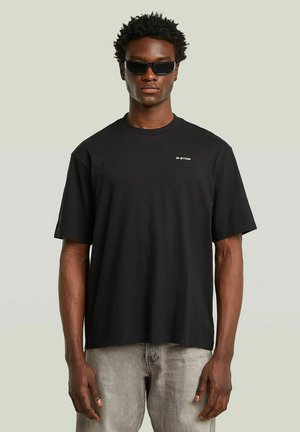 Uomo che indossa una maglietta nera G-Star, jeans grigi chiari e occhiali da sole neri rettangolari, in piedi contro uno sfondo chiaro e uniforme.