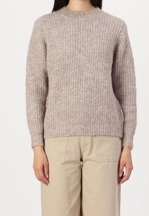 Personne portant un pull en tricot côtelé marron clair et un pantalon beige, debout devant un fond blanc uni.