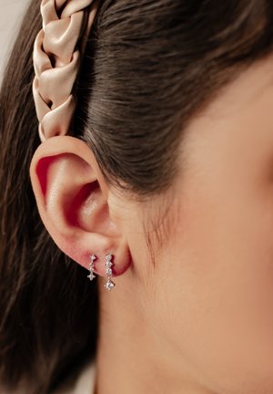 Silberne Tropfohrringe mit mehreren kleinen diamantenähnlichen Kristallen. Glatte Oberfläche mit minimalistischer Gestaltung. Die Haare sind zurückgesteckt.