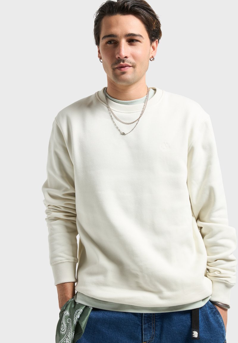 Sweatshirt crème avec col rond, manches longues et poignets côtelés. Associé à un t-shirt vert clair en dessous et un bandana vert à motif cachemire.