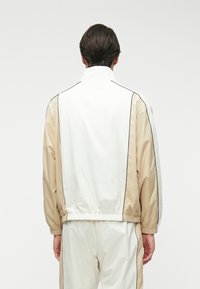 Ελαφρύ μπουφάν σε λευκό και μπεζ, με ψηλό γιακά, ελαστικούς καρπούς και πλευρικές ρίγες. Απαλή υφή και αθλητικός σχεδιασμός.