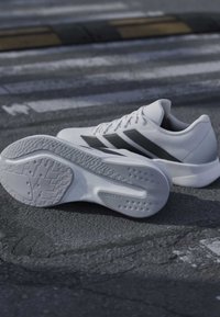 Chaussures de sport blanches à rayures noires posées sur l'asphalte près d'un passage piéton et d'un ralentisseur sous la lumière naturelle.