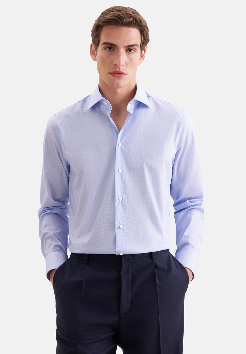 Jeune homme aux cheveux bruns portant une chemise bleu clair boutonnée et un pantalon foncé, debout les mains dans les poches devant un fond uni.