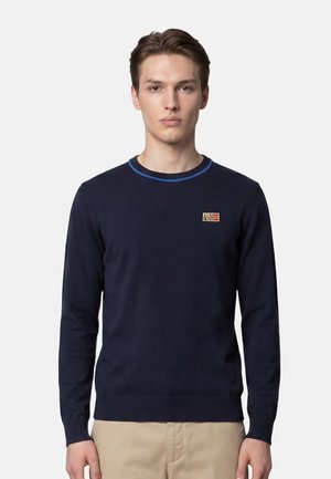 Giovane uomo indossa un maglione blu navy con una piccola toppa rettangolare sul petto e pantaloni beige, in piedi davanti a uno sfondo semplice.