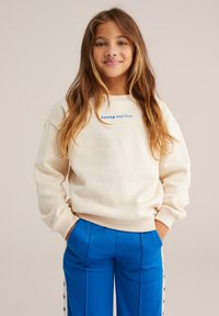 Krems krāsas džemperis ar zilu uzrakstu "Jauns un brīvs", ribotām piedurknēm un brīvu griezumu. Saskaņots ar zilām biksēm ar sānu zvaigžņu akcentiem.