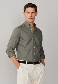 Mężczyzna w oliwkowo-zielonej koszuli na guziki z podwiniętymi rękawami, białych spodniach i ciemnym pasku, stoi z rękami w kieszeniach na szarym tle.