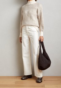 Beige flauschiger Pullover mit hohem Kragen, weiße gerade Jeans, dunkel gesteppte Tasche und dunkel glänzende Schuhe.