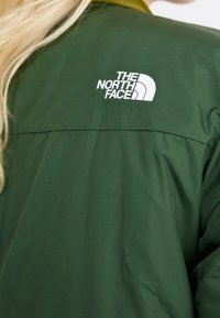 Personne blonde portant une veste vert foncé avec un logo blanc The North Face sur le haut du dos.
