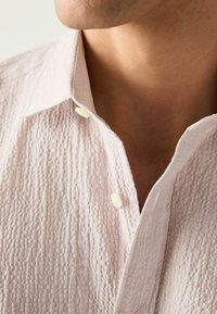 Světle růžová texturovaná košile s mírným vaflovým vzorem, se standartním límcem a dvěma bílými knoflíky u krku.