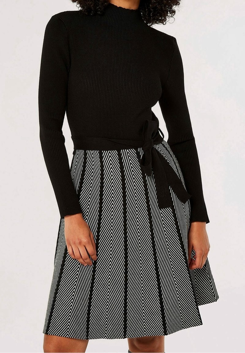 Femme portant un haut noir à manches longues côtelé et une jupe à motif chevron noir et blanc avec une ceinture noire nouée à la taille.