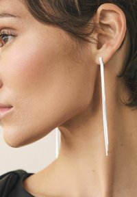 Lang sølvøreringe med et slankt, forlænget design, der hænger lige under øreflippen og har en glat, metallisk overflade.