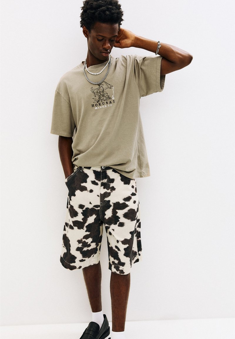 Volné olivově zelené tričko s grafickým potiskem, kombinované s šortkami s černobílým vzorem kravské kůže. Doplněno o vrstvené náhrdelníky a nazouvací boty.