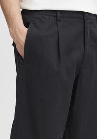 Pantalones negros hechos de tela ligera que presentan pliegues delanteros, bolsillos laterales y un ajuste ligeramente holgado con una textura suave.