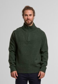 Donkergroene wollen trui met hoge geribde kraag, halve rits aan de voorkant en geribde manchetten, met een textuur en zachte afwerking.