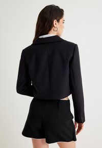 Geknipte zwarte blazer met een gelapte kraag, lange mouwen en knopenaccenten op de manchetten. Draagt over een wit shirt en gecombineerd met zwarte shorts.