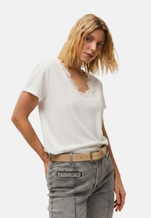 Mujer con cabello rubio que lleva una camiseta blanca con escote en V y ribete de encaje, pantalones grises de cintura alta y un cinturón beige, de pie con una mano en el bolsillo.