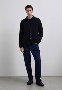 Jeune homme debout portant un cardigan boutonné texturé foncé, un pantalon marine et des chaussures noires, sur un fond gris clair uni.