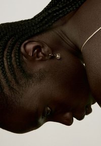 Pendiente de oro en forma de oreja con un diseño de ojo y acentos espigados. El modelo tiene el cabello trenzado y su tono de piel es rico y profundo. Se muestra en vista de perfil.