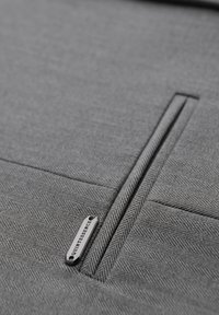 Gros plan d'une poche en tissu chevrons gris avec une fente verticale et une étiquette en métal portant l'inscription « QUINTESSENCE ».
