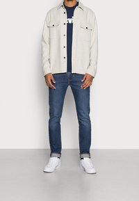 Ljusgrå skjorta med knappar och två bröstfickor över en mörkblå t-shirt. Matchad med snäva blå jeans och vita sneakers.