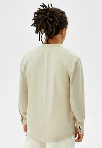 Beige overhemd met lange mouwen gemaakt van lichtgewicht stof, met een kraag, een detail van een rugpand en manchetten met knopen. De textuur oogt zacht en licht getextureerd.